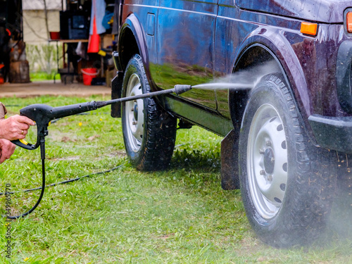 Car wash in the afternoon outdoors with a high-pressure apparatus. A powerful spray with water splashes away the dirt from the purple body of the car, from glass, wheels and tires.