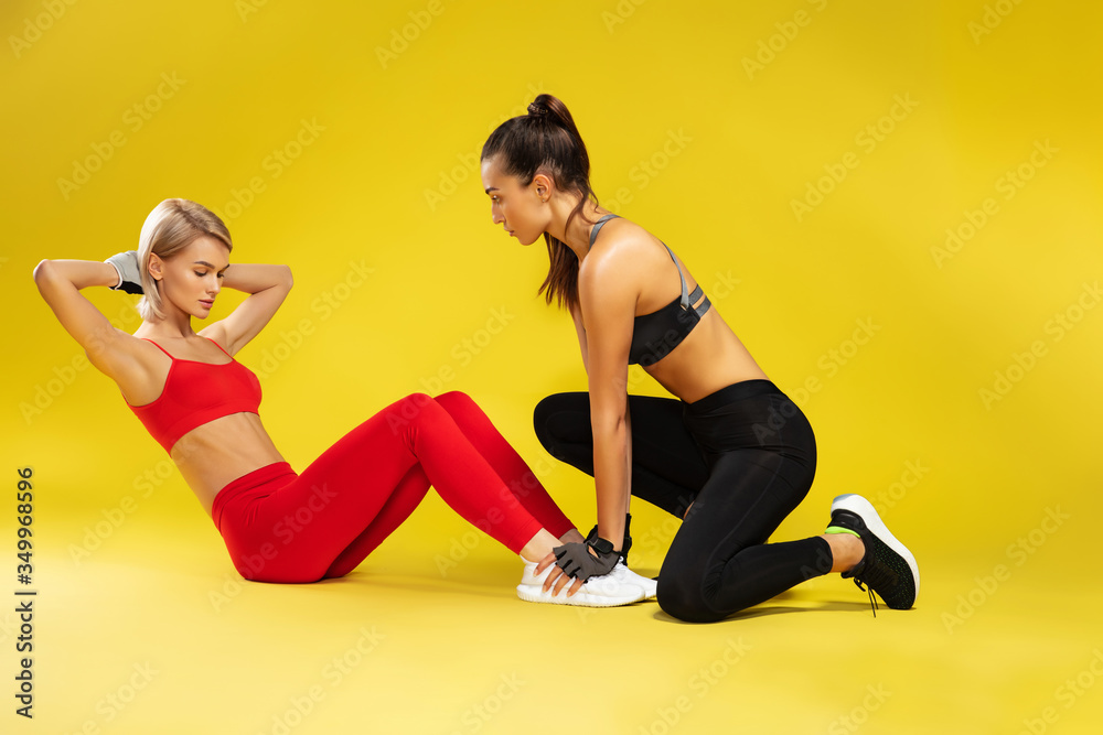 Pumping press. Attractive sporty blonde woman in red sportswear exercising with personal trainer against yellow bakground. Fitness girls