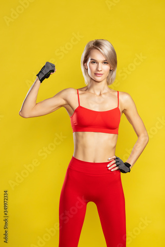 Fototapeta Naklejka Na Ścianę i Meble -  Strong arms. Young beautiful blonde woman in red sportswear showing her biceps and looking at camera while standing against yellow background