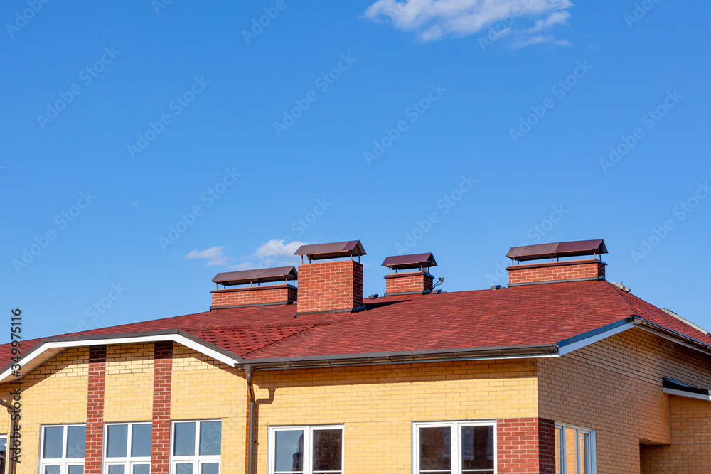 A cottage roof with slopes, tides, and a chimney against a blue sky. Brown roof of bituminous tiles for design on the theme of roofing, construction, architecture.
