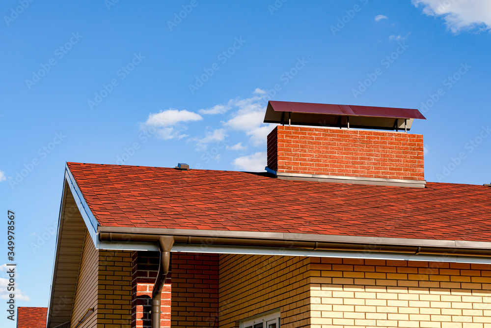 A cottage roof with slopes, tides, and a chimney against a blue sky. Brown roof of bituminous tiles for design on the theme of roofing, construction, architecture. Copy space