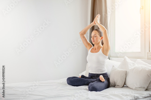 calm caucasian yogi woman sit on bed with crossed legs and hands up , meditate in the morning during quarantine, wearing sportswear isolated in light bedroom