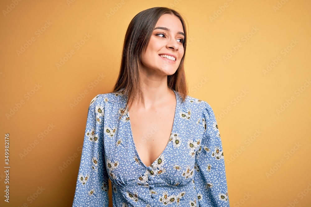 Young beautiful brunette woman wearing casual floral t-shirt standing over yellow background looking away to side with smile on face, natural expression. Laughing confident. Young beautiful brunette woman wearing casual floral t-shirt standing over yellow background looking away to side with smile on face, natural expression. Laughing confident.