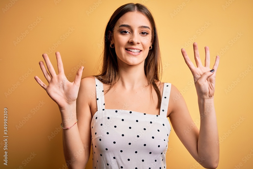 Young beautiful brunette woman wearing casual dress standing over yellow background showing and pointing up with fingers number nine while smiling confident and happy. Young beautiful brunette woman wearing casual dress standing over yellow background showing and pointing up with fingers number nine while smiling confident and happy.