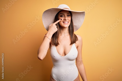 Young beautiful brunette woman on vacation wearing swimsuit and summer hat Pointing with hand finger to face and nose, smiling cheerful. Beauty concept