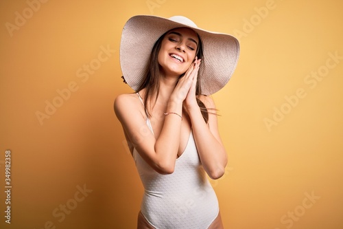 Young beautiful brunette woman on vacation wearing swimsuit and summer hat sleeping tired dreaming and posing with hands together while smiling with closed eyes.