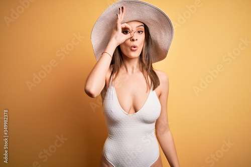 Young beautiful brunette woman on vacation wearing swimsuit and summer hat doing ok gesture shocked with surprised face, eye looking through fingers. Unbelieving expression.