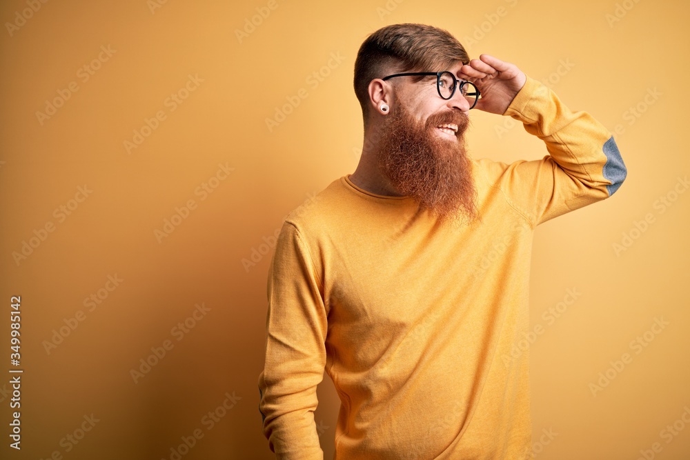 Handsome Irish redhead man with beard wearing glasses over yellow isolated background very happy and smiling looking far away with hand over head. Searching concept. Handsome Irish redhead man with beard wearing glasses over yellow isolated background very happy and smiling looking far away with hand over head. Searching concept.