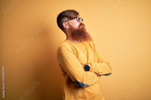 Handsome Irish redhead man with beard wearing glasses over yellow isolated background looking to the side with arms crossed convinced and confident