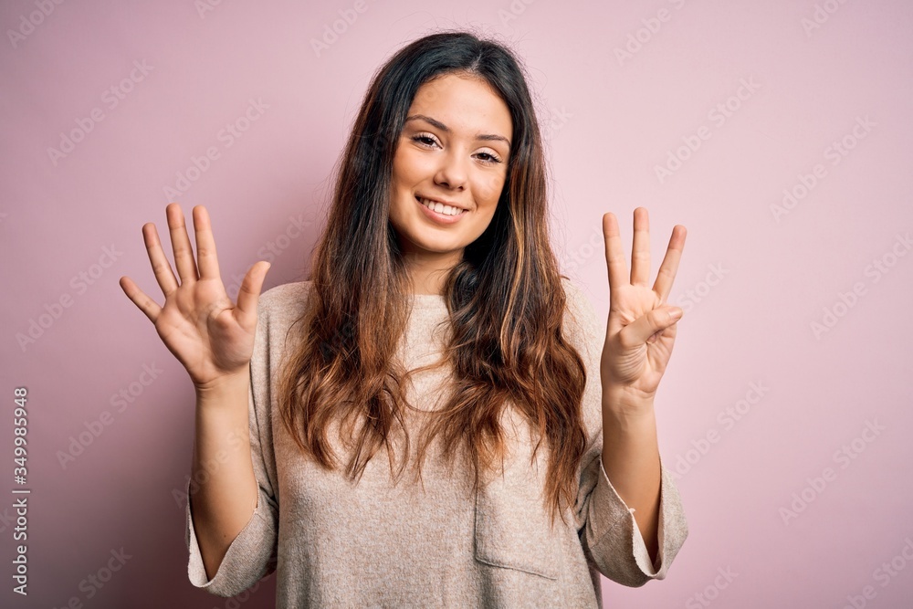 Young beautiful brunette woman wearing casual sweater standing over pink background showing and pointing up with fingers number eight while smiling confident and happy. Young beautiful brunette woman wearing casual sweater standing over pink background showing and pointing up with fingers number eight while smiling confident and happy.