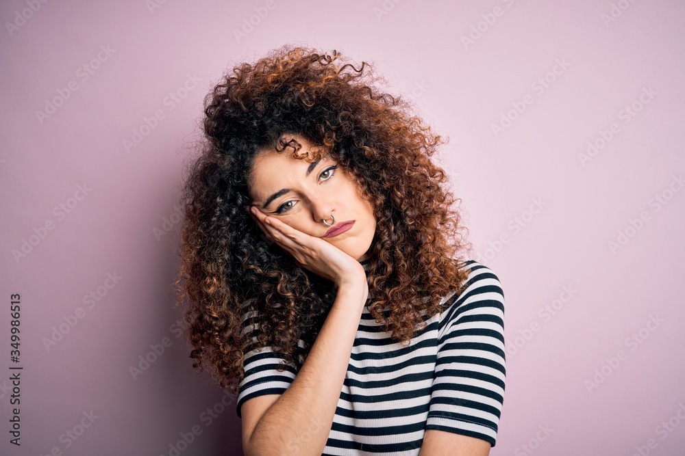 Young beautiful woman with curly hair and piercing wearing casual striped t-shirt thinking looking tired and bored with depression problems with crossed arms.