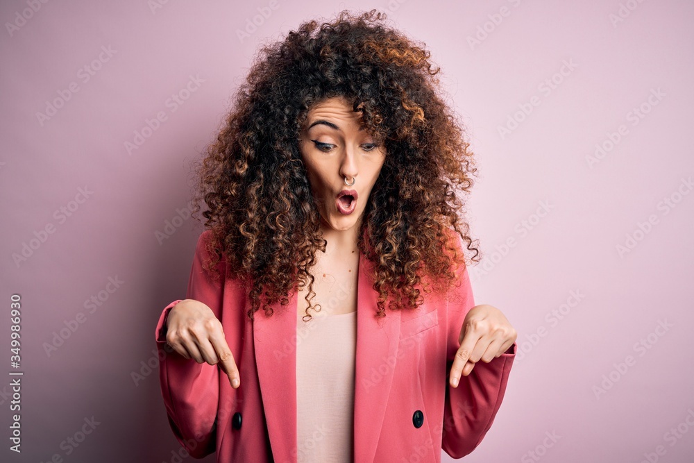 Young beautiful businesswoman with curly hair and piercing wearing elegant jacket Pointing down with fingers showing advertisement, surprised face and open mouth Young beautiful businesswoman with curly hair and piercing wearing elegant jacket Pointing down with fingers showing advertisement, surprised face and open mouth