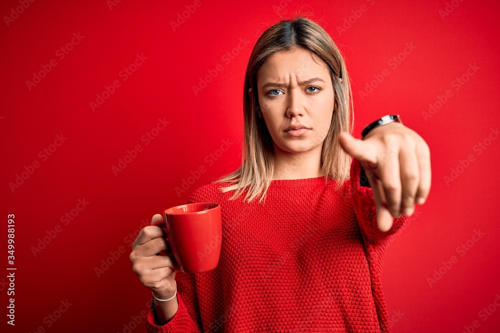 Naklejka premium Young beautiful woman drinking cup of coffee standing over isolated red background pointing with finger to the camera and to you, hand sign, positive and confident gesture from the front