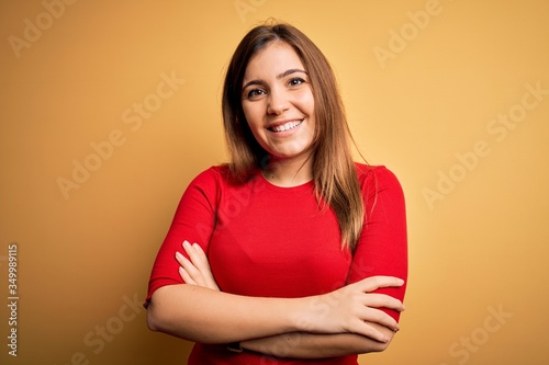 Beautiful young woman wearing casual red t-shirt over yellow isolated background happy face smiling with crossed arms looking at the camera. Positive person.