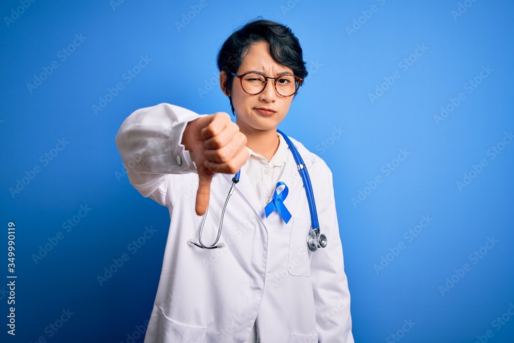 Young beautiful asian doctor girl wearing stethoscope and coat with blue cancer ribbon looking unhappy and angry showing rejection and negative with thumbs down gesture. Bad expression. Young beautiful asian doctor girl wearing stethoscope and coat with blue cancer ribbon looking unhappy and angry showing rejection and negative with thumbs down gesture. Bad expression.