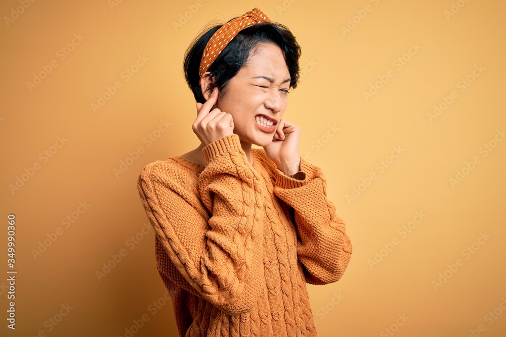 Young beautiful asian girl wearing casual sweater and diadem standing over yellow background covering ears with fingers with annoyed expression for the noise of loud music. Deaf concept. Young beautiful asian girl wearing casual sweater and diadem standing over yellow background covering ears with fingers with annoyed expression for the noise of loud music. Deaf concept.