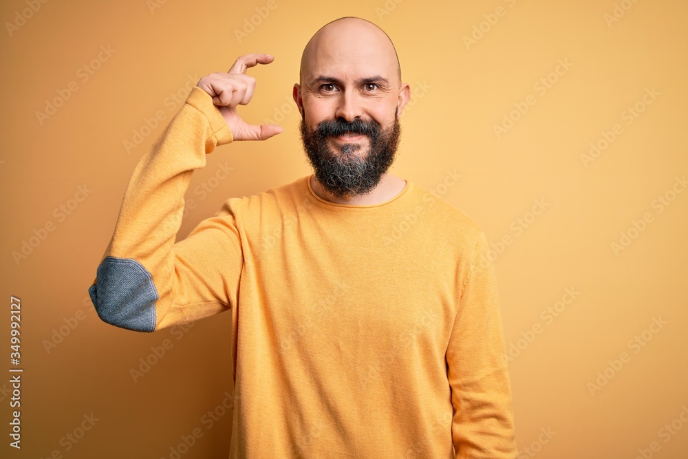 Handsome bald man with beard wearing casual sweater standing over yellow background smiling and confident gesturing with hand doing small size sign with fingers looking and the camera. Measure concept Handsome bald man with beard wearing casual sweater standing over yellow background smiling and confident gesturing with hand doing small size sign with fingers looking and the camera. Measure concept