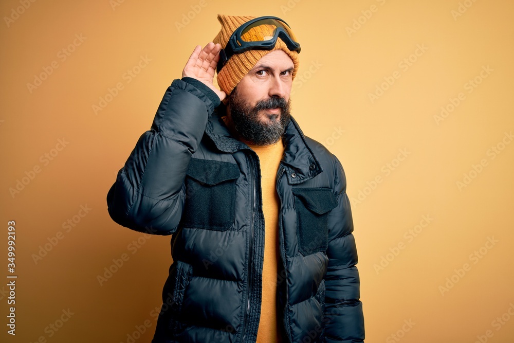 Handsome skier bald man with beard skiing wearing snow sportswear and ski goggles smiling with hand over ear listening an hearing to rumor or gossip. Deafness concept. Handsome skier bald man with beard skiing wearing snow sportswear and ski goggles smiling with hand over ear listening an hearing to rumor or gossip. Deafness concept.