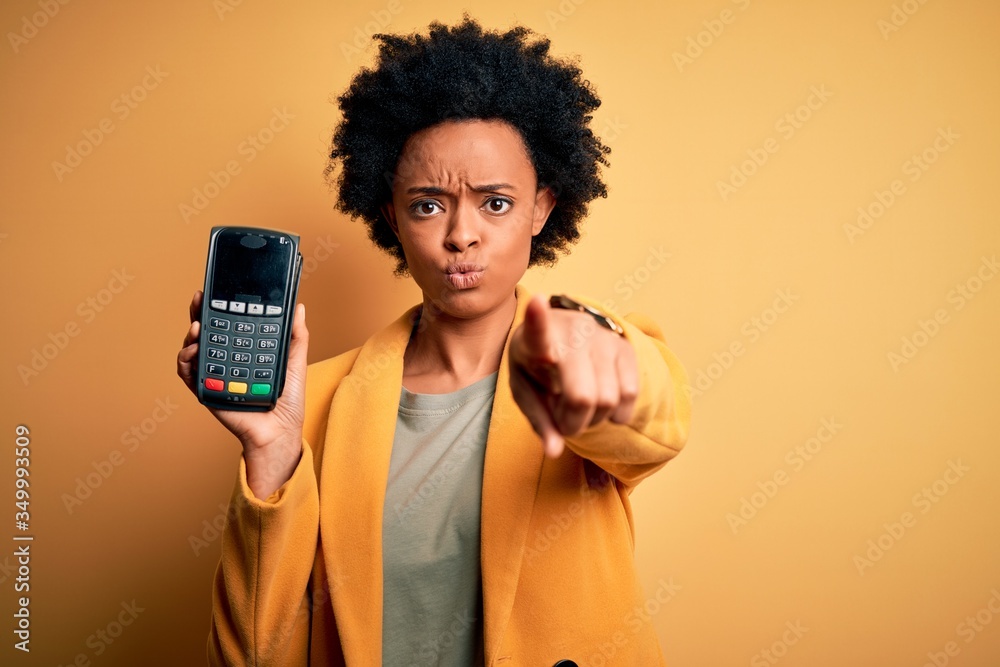 Young African American afro woman with curly hair holding dataphone payment device pointing with finger to the camera and to you, hand sign, positive and confident gesture from the front Young African American afro woman with curly hair holding dataphone payment device pointing with finger to the camera and to you, hand sign, positive and confident gesture from the front