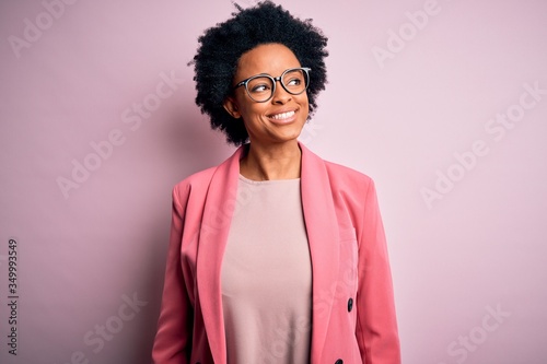 Young beautiful African American afro businesswoman with curly hair wearing pink jacket smiling looking to the side and staring away thinking.