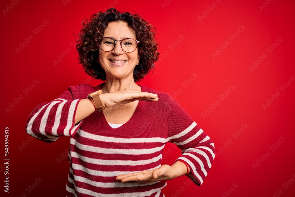 Middle age beautiful curly hair woman wearing casual striped sweater over red background gesturing with hands showing big and large size sign, measure symbol. Smiling looking at the camera. Measuring Middle age beautiful curly hair woman wearing casual striped sweater over red background gesturing with hands showing big and large size sign, measure symbol. Smiling looking at the camera. Measuring
