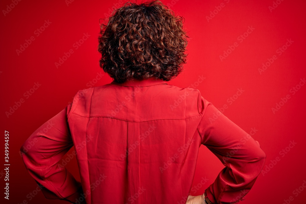 Middle age beautiful curly hair woman wearing casual shirt and glasses over red background standing backwards looking away with arms on body Middle age beautiful curly hair woman wearing casual shirt and glasses over red background standing backwards looking away with arms on body