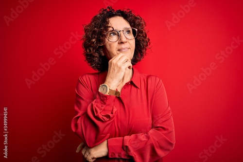 Middle age beautiful curly hair woman wearing casual shirt and glasses over red background with hand on chin thinking about question  pensive expression. Smiling with thoughtful face. Doubt concept.