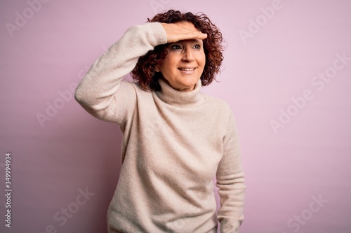 Middle age beautiful curly hair woman wearing casual turtleneck sweater over pink background very happy and smiling looking far away with hand over head. Searching concept. © Krakenimages.com