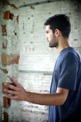 young man leaning against wall, a young man in blue t-shirt and gray shorts stands in an abandoned and ruined building