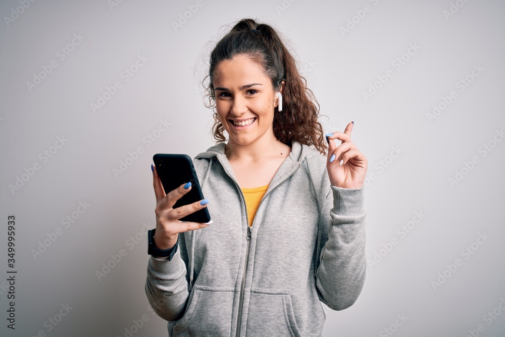 Fototapeta premium Beautiful sportswoman with curly hair listening to music using smartphone and earphones surprised with an idea or question pointing finger with happy face, number one