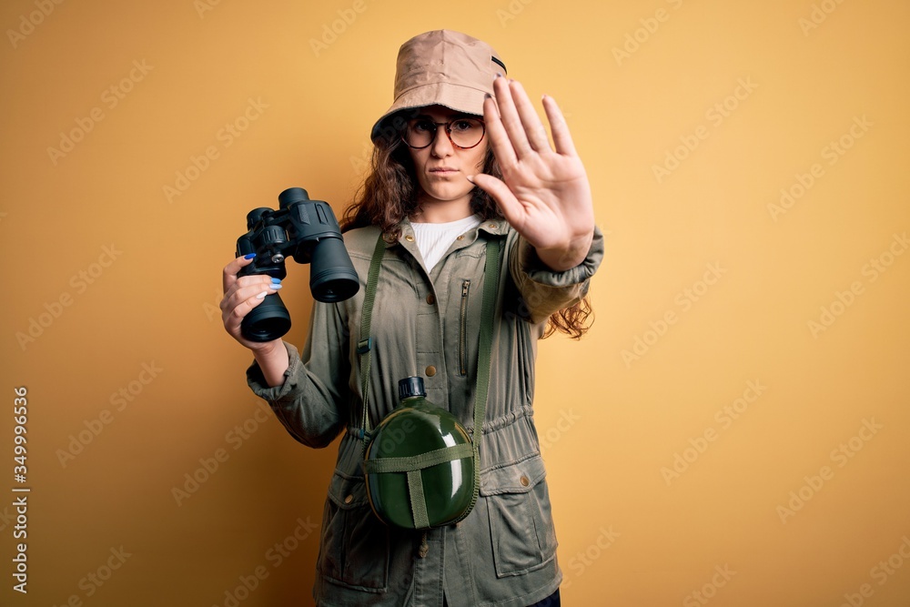 Beautiful tourist woman on vacation wearing explorer hat and water canteen using binoculars with open hand doing stop sign with serious and confident expression, defense gesture