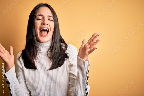 Young brunette woman with blue eyes wearing casual sweater over isolated yellow background celebrating mad and crazy for success with arms raised and closed eyes screaming excited. Winner concept
