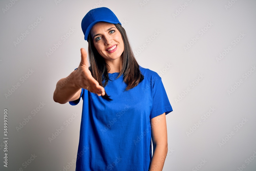 Young delivery woman with blue eyes wearing cap standing over blue background smiling friendly offering handshake as greeting and welcoming. Successful business.