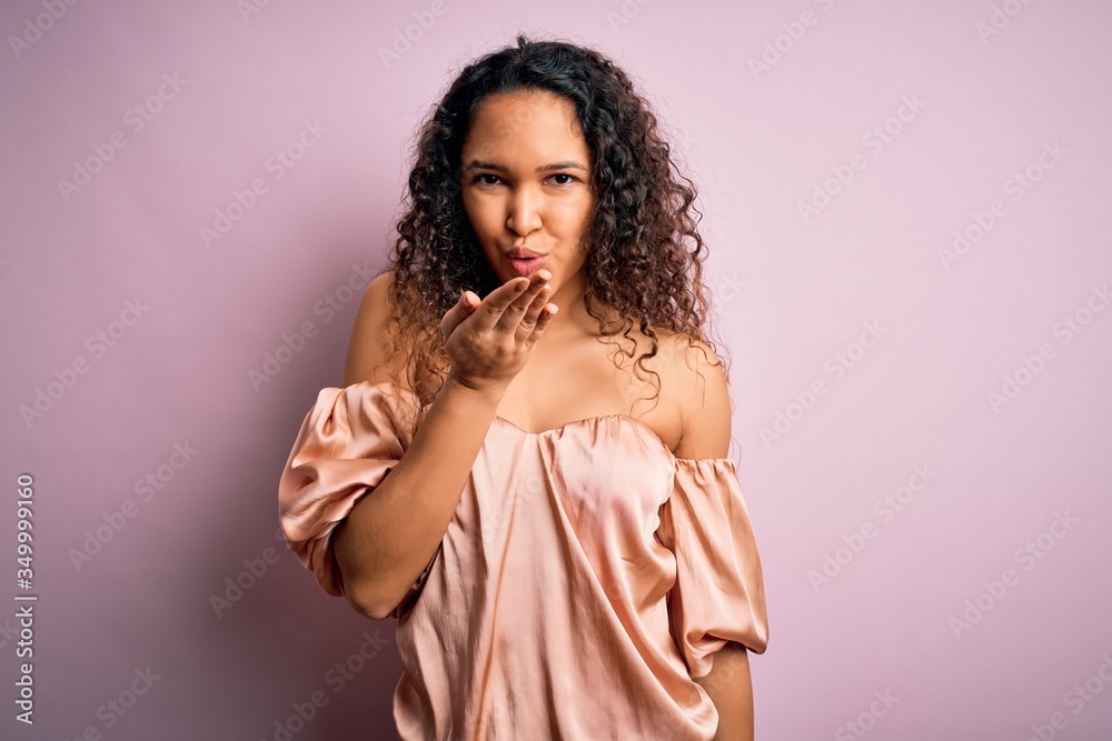 Young beautiful woman with curly hair wearing casual t-shirt standing over pink background looking at the camera blowing a kiss with hand on air being lovely and sexy. Love expression. Young beautiful woman with curly hair wearing casual t-shirt standing over pink background looking at the camera blowing a kiss with hand on air being lovely and sexy. Love expression.