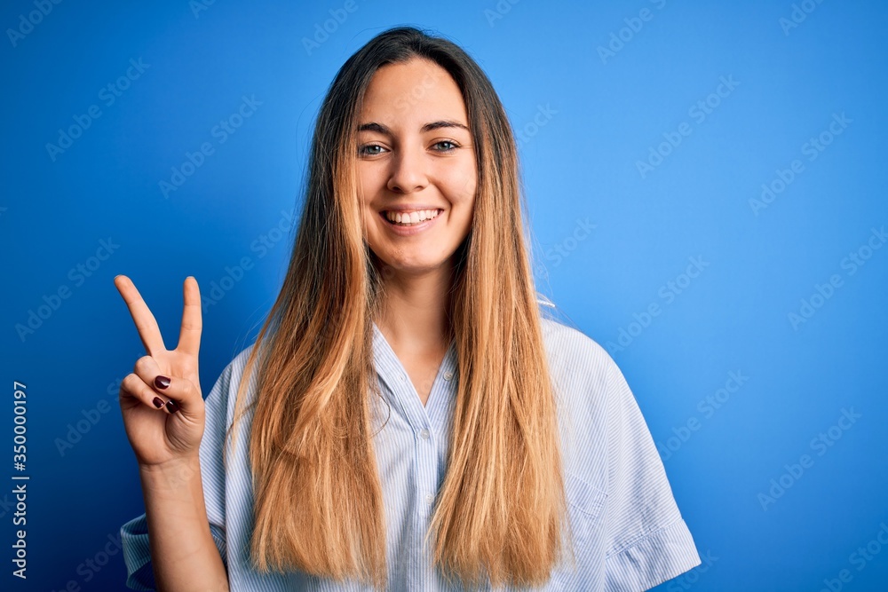 Young beautiful blonde woman with blue eyes wearing striped shirt over blue background smiling with happy face winking at the camera doing victory sign. Number two. Young beautiful blonde woman with blue eyes wearing striped shirt over blue background smiling with happy face winking at the camera doing victory sign. Number two.