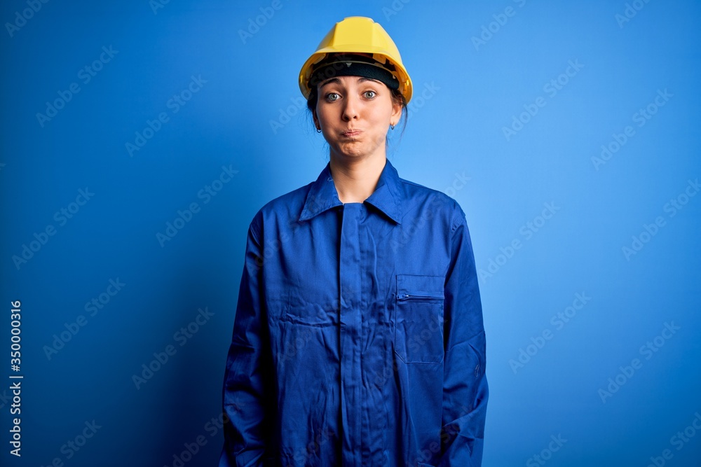 Young beautiful worker woman with blue eyes wearing security helmet and uniform puffing cheeks with funny face. Mouth inflated with air, crazy expression. Young beautiful worker woman with blue eyes wearing security helmet and uniform puffing cheeks with funny face. Mouth inflated with air, crazy expression.