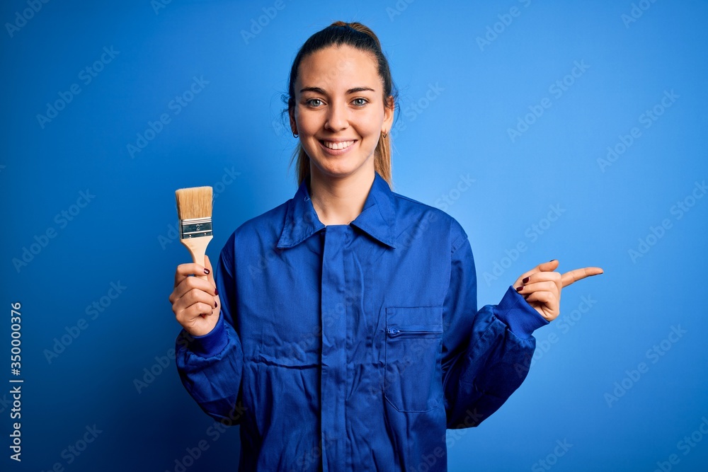 Young beautiful painter woman with blue eyes painting wearing uniform using paint brush very happy pointing with hand and finger to the side