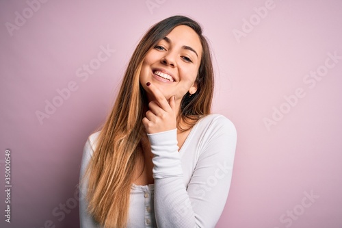 Young beautiful blonde woman with blue eyes wearing white t-shirt over pink background looking confident at the camera smiling with crossed arms and hand raised on chin. Thinking positive.