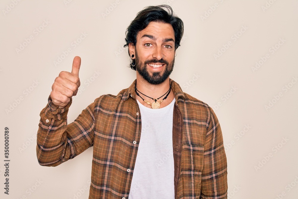 Young handsome hispanic bohemian man wearing hippie style standing over isolated background doing happy thumbs up gesture with hand. Approving expression looking at the camera showing success. Young handsome hispanic bohemian man wearing hippie style standing over isolated background doing happy thumbs up gesture with hand. Approving expression looking at the camera showing success.