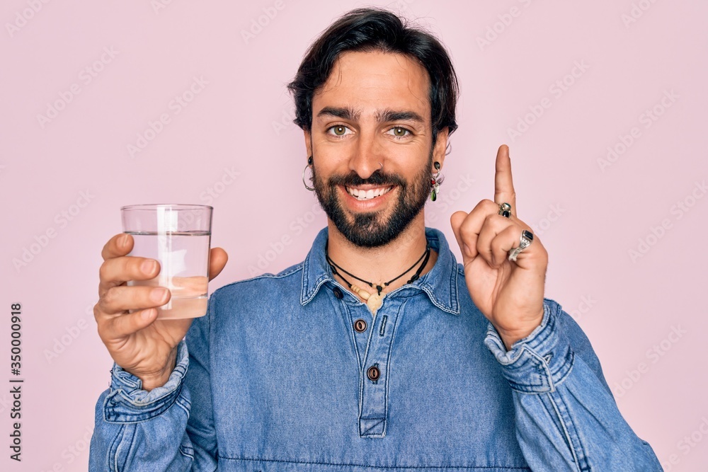 Young handsome hispanic bohemian hipster man with bear drinking a fresh glass of water surprised with an idea or question pointing finger with happy face, number one Young handsome hispanic bohemian hipster man with bear drinking a fresh glass of water surprised with an idea or question pointing finger with happy face, number one