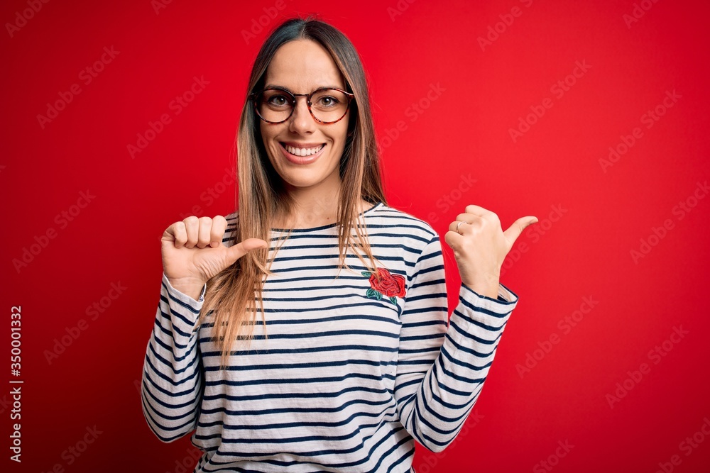 Young beautiful blonde woman with blue eyes wearing glasses standing over red background Pointing to the back behind with hand and thumbs up, smiling confident Young beautiful blonde woman with blue eyes wearing glasses standing over red background Pointing to the back behind with hand and thumbs up, smiling confident