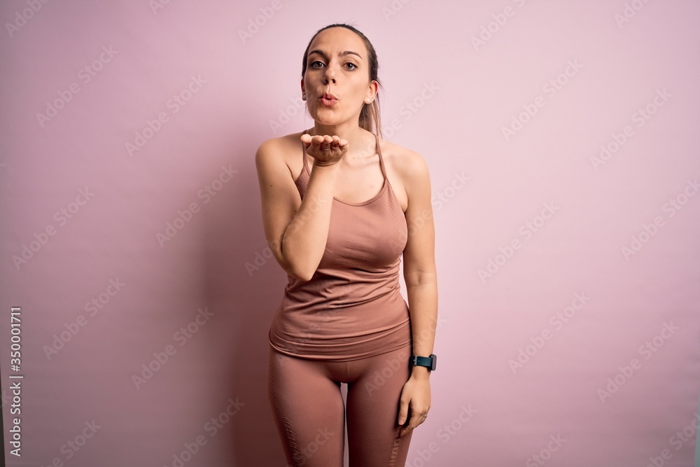 Naklejka premium Young blonde fitness woman wearing sport workout clothes over pink isolated background looking at the camera blowing a kiss with hand on air being lovely and sexy. Love expression.