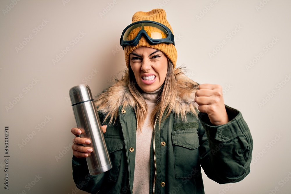 Young beautiful brunette skier woman wearing ski goggles drinking thermo with coffe annoyed and frustrated shouting with anger, crazy and yelling with raised hand, anger concept