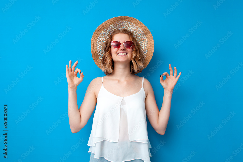 girl in a sun hat and glasses shows okay gesture on a blue isolated background, joyful woman in summer clothes, summer concept