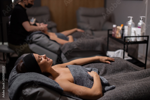 side view on calm relaxed caucasian woman lying in wellbeing salon before skin treatment, woman get face lift massage in the background