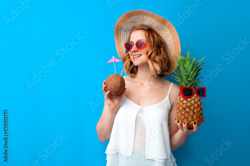 girl with coconut cocktail and pineapple in a sun hat and glasses on a blue background, woman with a tropical cocktail on vacation, summer concept