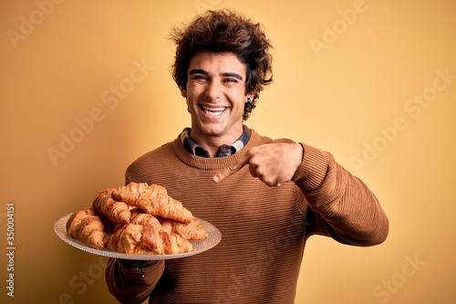 Young handsome man holding plate with croissants standing over isolated yellow background very happy pointing with hand and finger