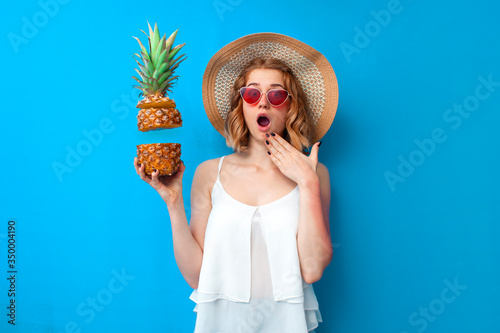 young shocked girl in summer clothes holds a sliced pineapple and wonders on a blue isolated background, woman with tropical fruit in summer
