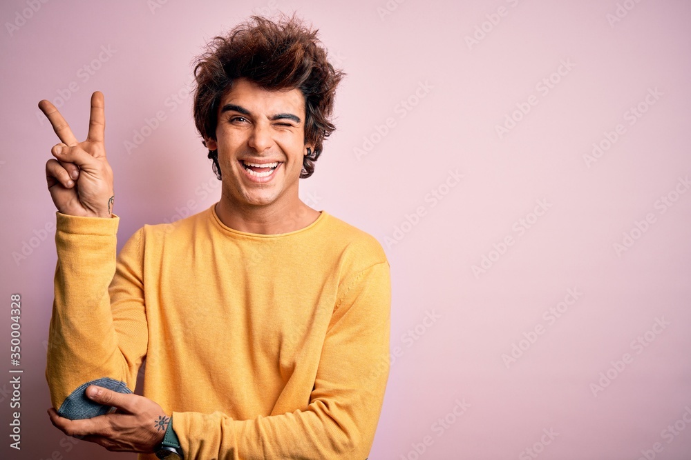 Young handsome man wearing yellow casual t-shirt standing over isolated pink background smiling with happy face winking at the camera doing victory sign. Number two. Young handsome man wearing yellow casual t-shirt standing over isolated pink background smiling with happy face winking at the camera doing victory sign. Number two.