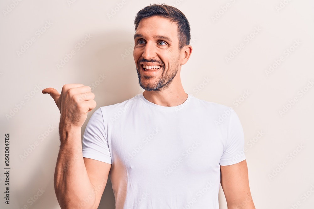 Young handsome man wearing casual t-shirt standing over isolated white background pointing thumb up to the side smiling happy with open mouth Young handsome man wearing casual t-shirt standing over isolated white background pointing thumb up to the side smiling happy with open mouth